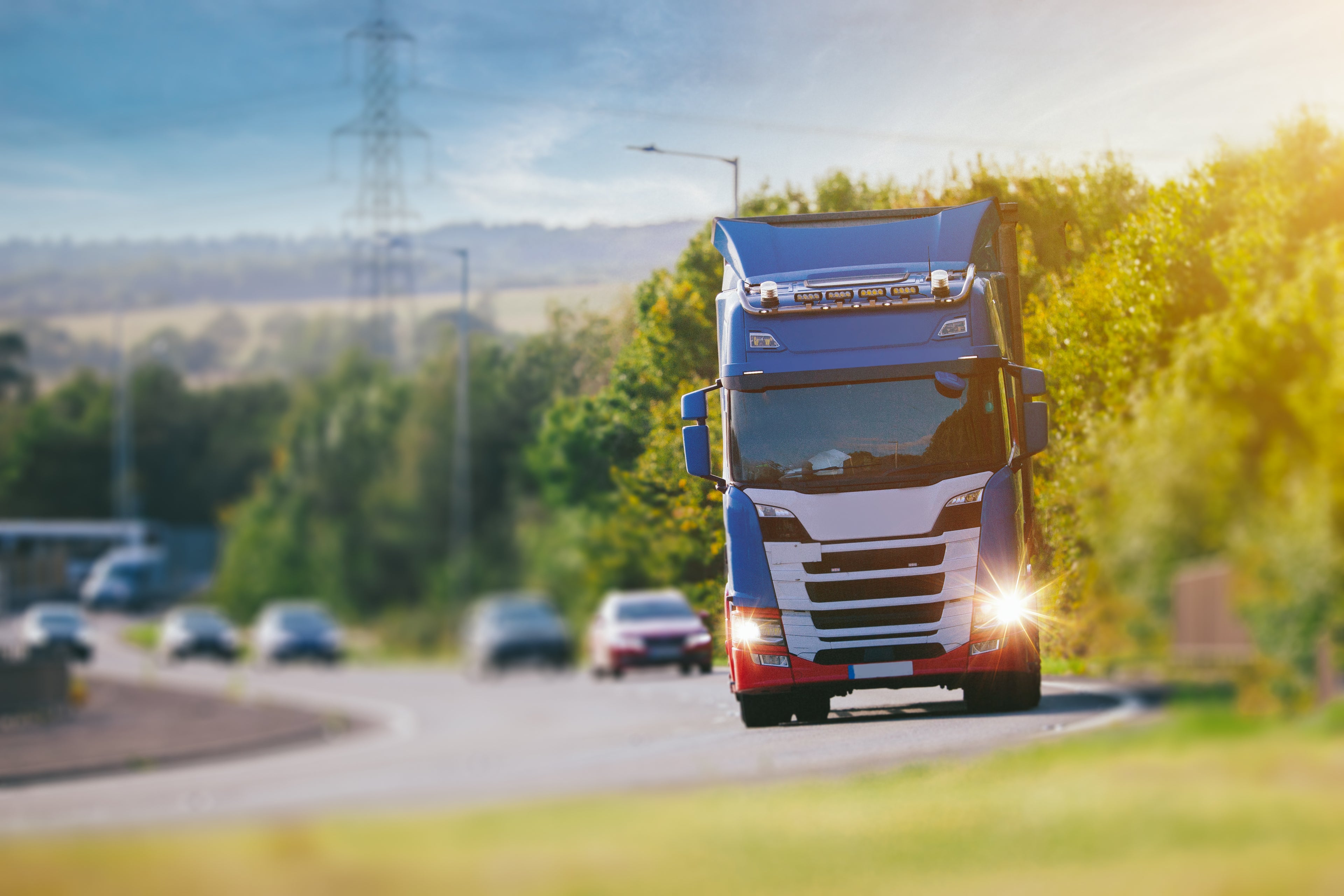 A blue and white Scania semi-truck driving on a curved road with trees and hills in the background under a bright sky.